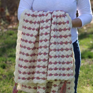 May include: A crocheted blanket with pink and white stripes and tassels. The blanket is folded in half and held by a person.