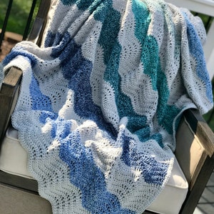 May include: A crocheted blanket in shades of blue and gray, draped over a chair. The blanket has a wavy pattern and is made with a soft yarn.