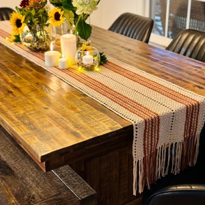 Könnte beinhalten: Ein Esstisch aus Holz mit einem gehäkelten Tischläufer in Beige und Orange mit Fransen. Der Tisch ist mit Kerzen und Sonnenblumen in Glasvasen gedeckt.