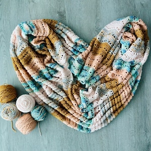 May include: A heart-shaped infinity scarf featuring a crochet pattern with alternating stripes of mustard yellow, soft pink, white, and variegated teal blue yarn, accompanied by five small matching yarn balls in the same colour palette.