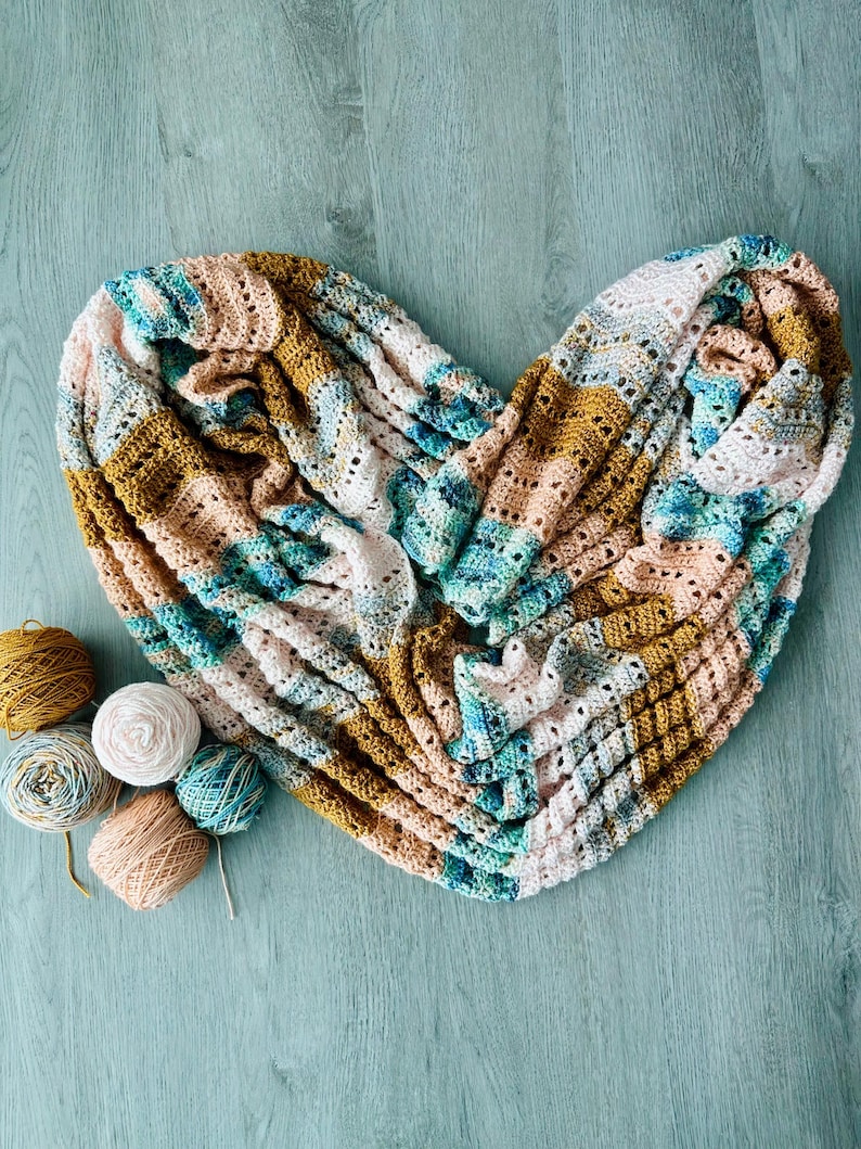 May include: A heart-shaped infinity scarf featuring a crochet pattern with alternating stripes of mustard yellow, soft pink, white, and variegated teal blue yarn, accompanied by five small matching yarn balls in the same colour palette.