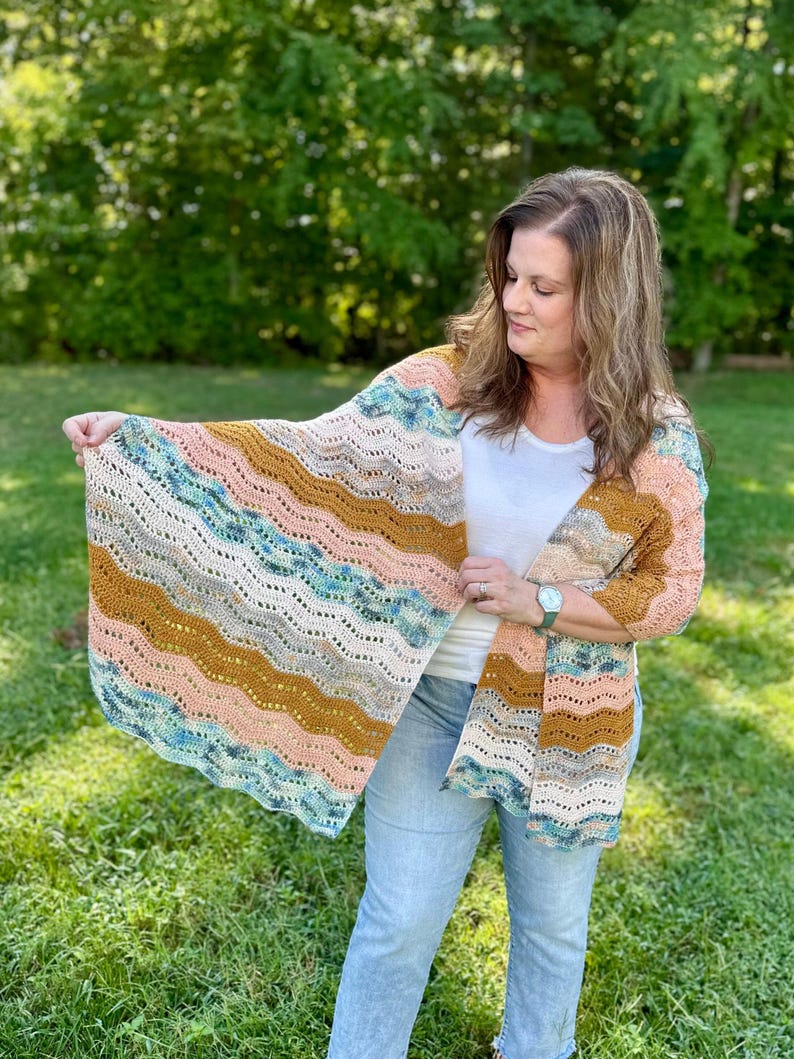 May include: A crochet shawl featuring a wavy, ripple stitch pattern in horizontal stripes of peach, mustard yellow, cream, and variegated blue and green yarn. The lightweight garment has an openwork texture and a scalloped edge design.