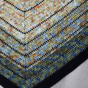May include: A handmade crochet shawl in a triangular shape. The shawl features a colour gradient from light to dark, with a black border. The colours include blue, green, orange, and black, creating a textured, patterned design.
