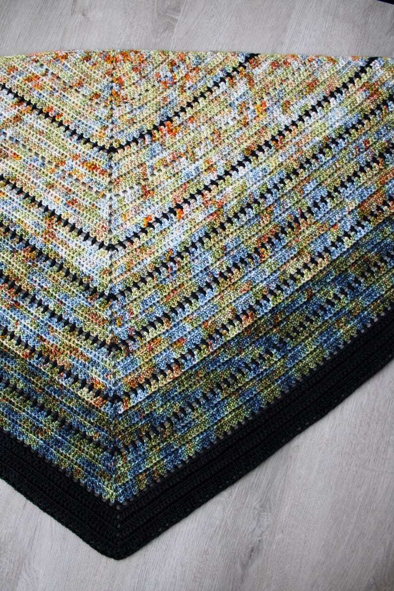 May include: A handmade crochet shawl in a triangular shape. The shawl features a colour gradient from light to dark, with a black border. The colours include blue, green, orange, and black, creating a textured, patterned design.