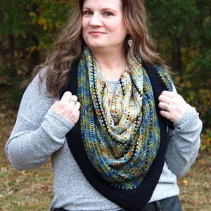 May include: A hand-knitted shawl with a black border. The shawl features a mix of blue, green, and orange colours. The woman in the image is wearing a grey long-sleeve top and black jeans.
