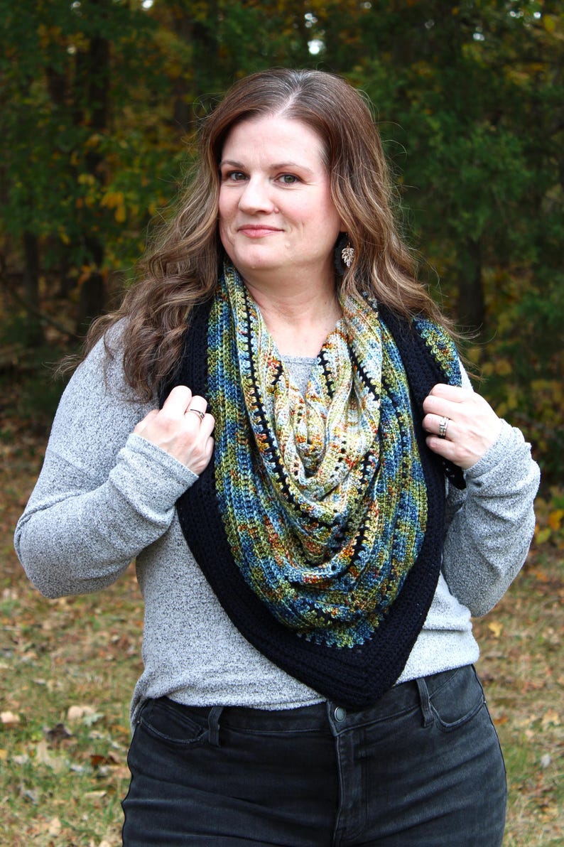 May include: A hand-knitted shawl with a black border. The shawl features a mix of blue, green, and orange colours. The woman in the image is wearing a grey long-sleeve top and black jeans.