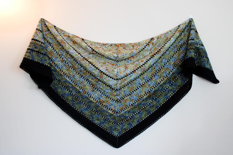 May include: A hand-knitted triangular shawl with a black border and a striped pattern. The shawl features a gradient of colours, including blue, green, and orange, and is made of soft yarn. The shawl is displayed against a white background.