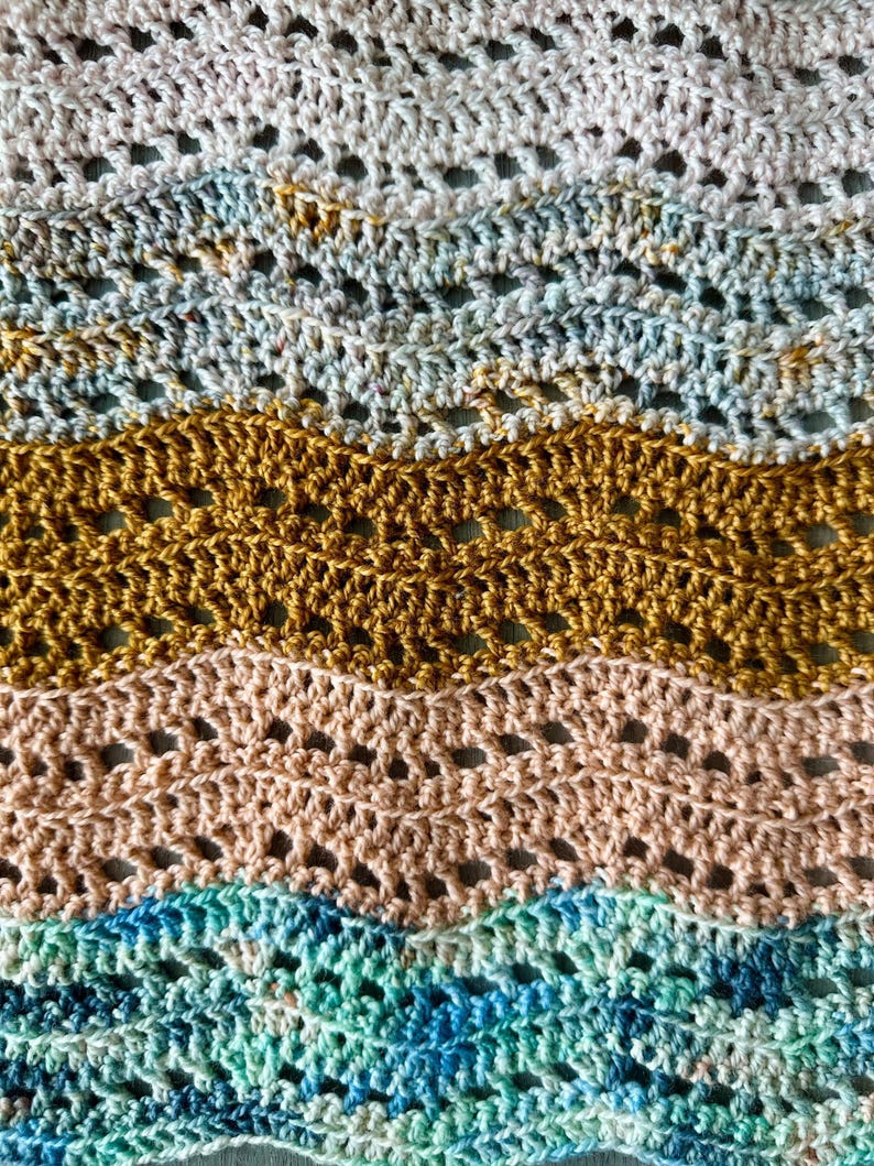May include: A crocheted fabric featuring a repeating wavy ripple pattern. The design consists of horizontal bands in alternating colours of cream, variegated blue and grey, solid mustard yellow, soft peach, and variegated teal and dark blue.