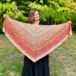 May include: A woman wearing a black dress holds a crocheted shawl with a gradient of orange, pink, and beige colors. The shawl has a scalloped edge.
