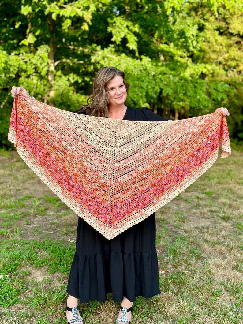 May include: A woman wearing a black dress holds a crocheted shawl with a gradient of orange, pink, and beige colors. The shawl has a scalloped edge.