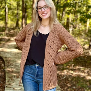 May include: A brown crochet cardigan with long sleeves. The open-front sweater is worn over a black top and blue jeans. The cardigan has a textured, open weave design. The background is a blurred view of a wooded area.