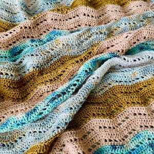 May include: A crocheted blanket featuring a wavy ripple pattern in shades of mustard yellow, soft peach, white, and variegated teal blue. A metallic copper-coloured crochet hook rests on the fabric, accompanied by a ball of matching yarn.