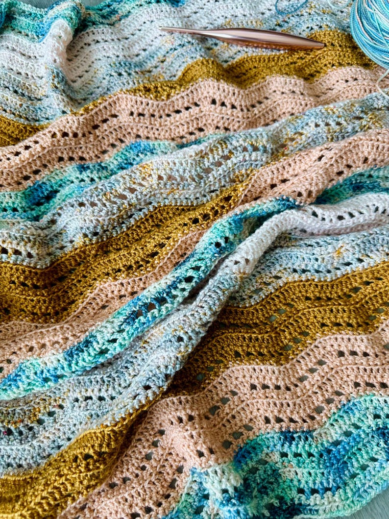 May include: A crocheted blanket featuring a wavy ripple pattern in shades of mustard yellow, soft peach, white, and variegated teal blue. A metallic copper-coloured crochet hook rests on the fabric, accompanied by a ball of matching yarn.