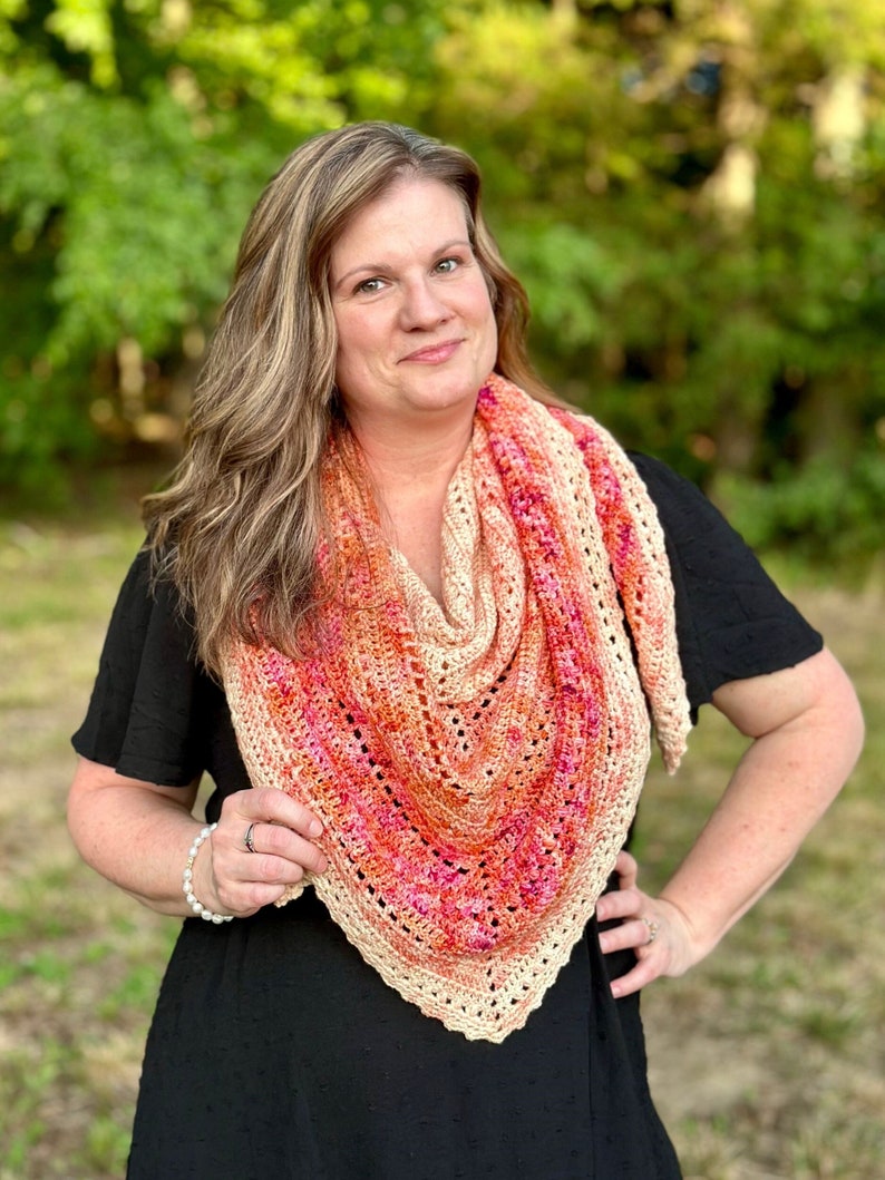 May include: A woman wearing a black dress with a crocheted shawl in shades of peach, orange, and pink.