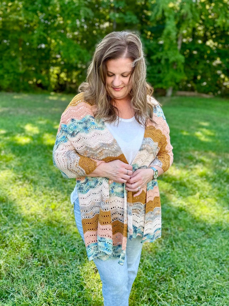 May include: A crochet cardigan featuring a wavy, multi-coloured pattern in shades of mustard yellow, soft peach, cream, and teal blue. The garment has an open front design with a textured, lacy stitch pattern throughout the fabric.