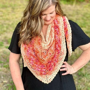 May include: A woman wearing a black dress with a crocheted shawl in shades of peach, pink, and orange. The shawl has a triangular shape with a delicate lace pattern.