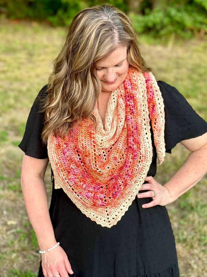 May include: A woman wearing a black dress with a crocheted shawl in shades of peach, pink, and orange. The shawl has a triangular shape with a delicate lace pattern.