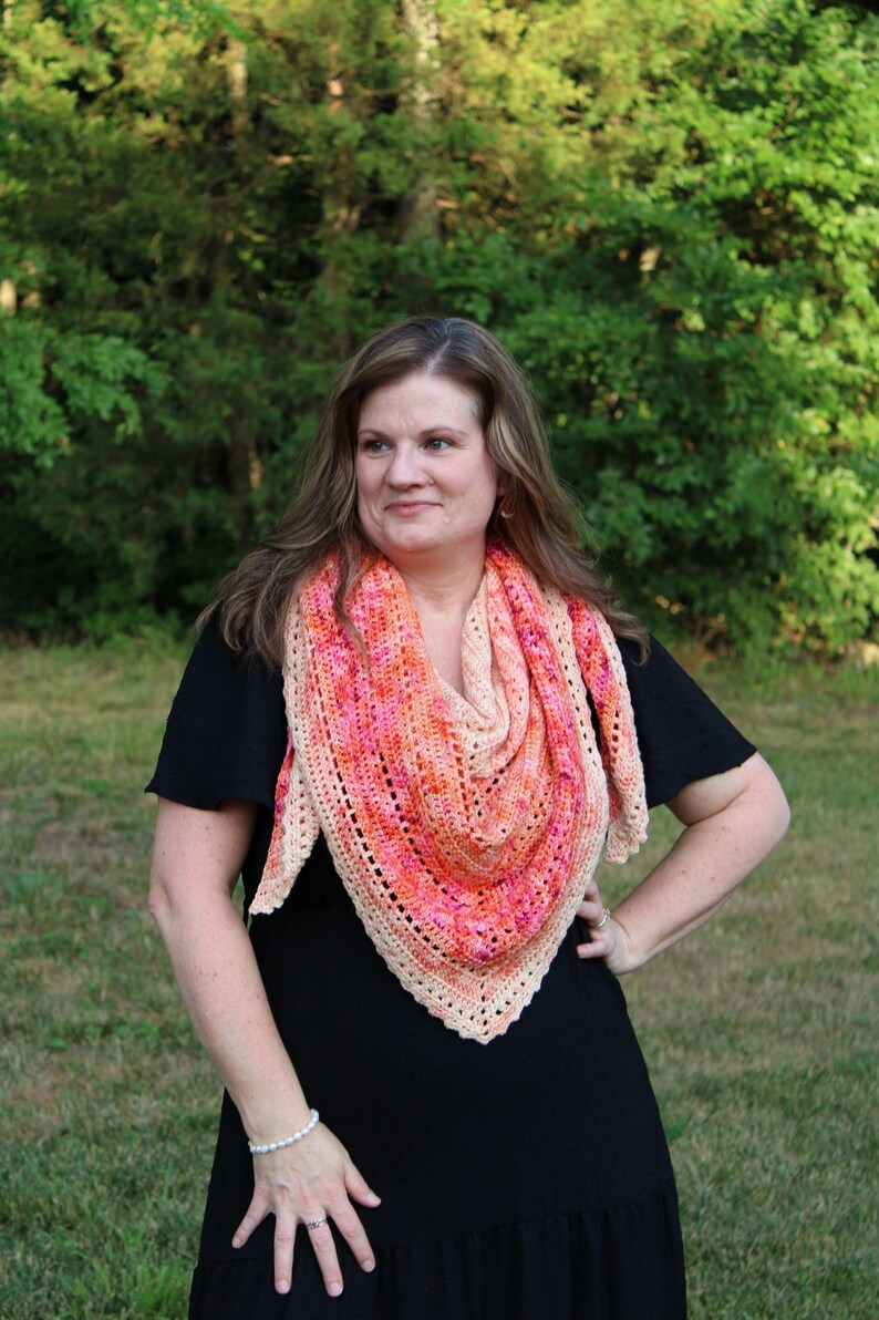 May include: A woman wearing a black dress with a crocheted shawl in shades of pink, orange, and cream. The shawl has a triangular shape and a delicate lace pattern.