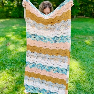 May include: A rectangular crochet blanket featuring a repeating wavy ripple pattern. The design incorporates horizontal stripes in shades of mustard yellow, soft peach, white, and a variegated blend of teal, blue, and grey yarn throughout the piece.