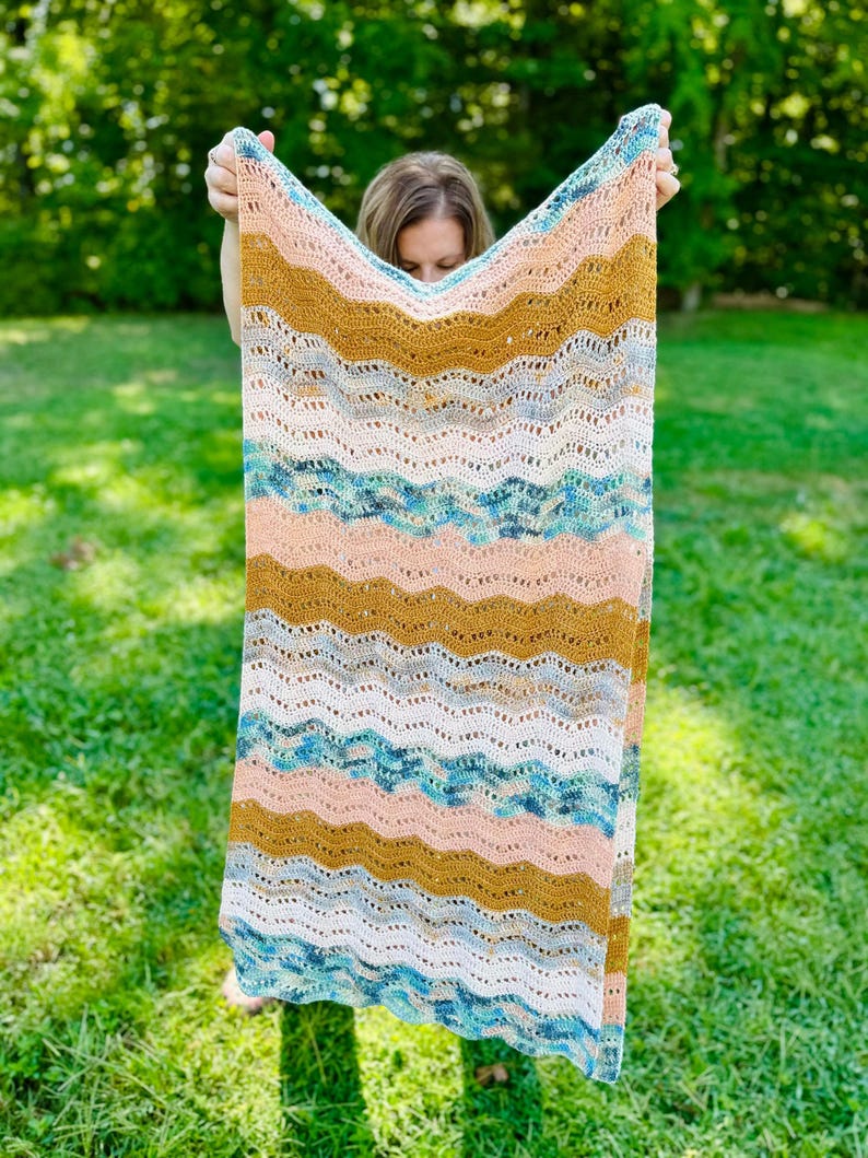 May include: A rectangular crochet blanket featuring a repeating wavy ripple pattern. The design incorporates horizontal stripes in shades of mustard yellow, soft peach, white, and a variegated blend of teal, blue, and grey yarn throughout the piece.