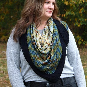May include: A colourful, crocheted shawl with a black border. The shawl features a mix of blue, green, and yellow yarn. The person is wearing a grey long-sleeved shirt and black jeans. The background is blurred with autumn foliage.