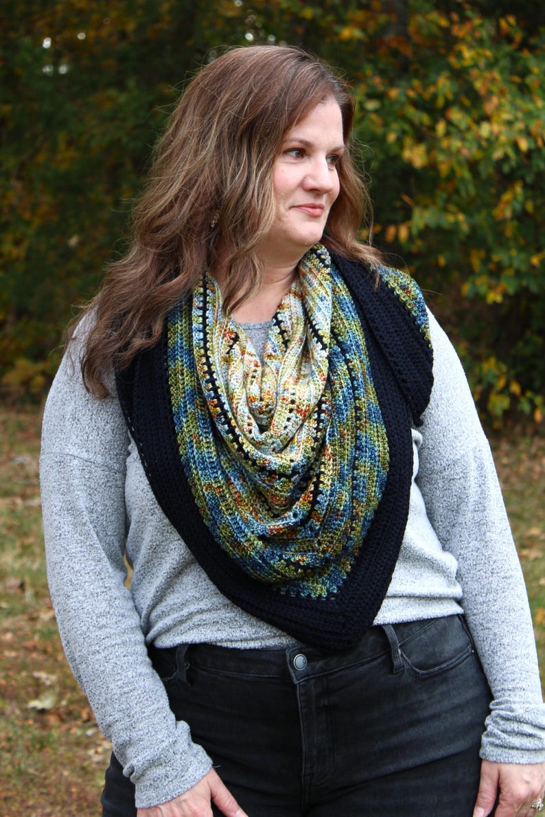 May include: A colourful, crocheted shawl with a black border. The shawl features a mix of blue, green, and yellow yarn. The person is wearing a grey long-sleeved shirt and black jeans. The background is blurred with autumn foliage.