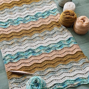 May include: A rectangular crochet blanket featuring a repeating wavy ripple pattern in shades of mustard yellow, teal blue, soft peach, and cream. A rose gold crochet hook rests on the fabric, accompanied by several matching yarn balls.