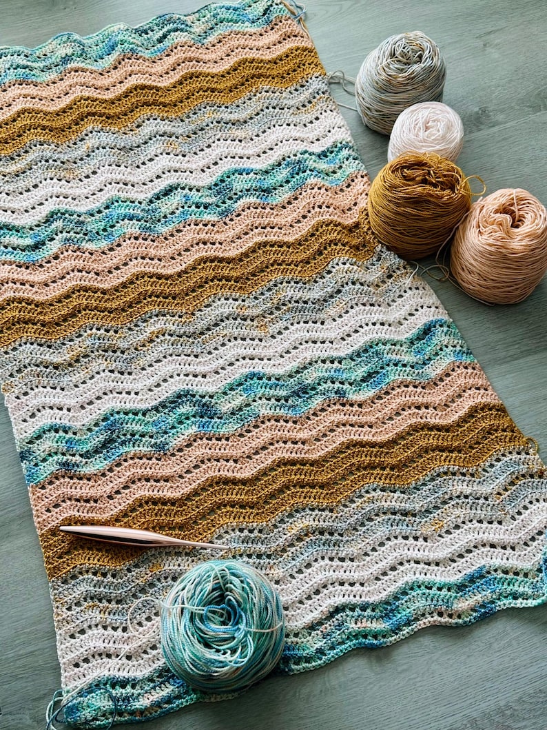May include: A rectangular crochet blanket featuring a repeating wavy ripple pattern in shades of mustard yellow, teal blue, soft peach, and cream. A rose gold crochet hook rests on the fabric, accompanied by several matching yarn balls.