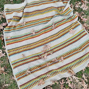 Op de afbeelding: Een handgemaakte gehaakte deken met een gestreept patroon in tinten grijs, rood, geel en blauwgroen. De deken ligt op gras en gevallen bladeren. De deken is ongeveer 152 cm breed en 178 cm lang.