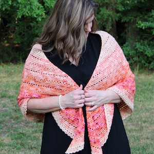 May include: A woman wearing a black dress with a crocheted shawl in shades of peach, pink, and white. The shawl has a scalloped edge and is draped around her shoulders.