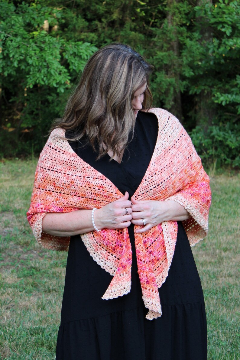 May include: A woman wearing a black dress with a crocheted shawl in shades of peach, pink, and white. The shawl has a scalloped edge and is draped around her shoulders.