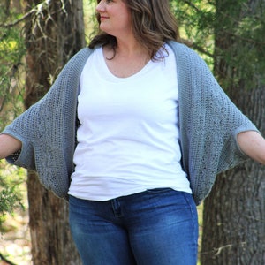 Może przedstawiać: Kobieta w szarej dzianinowej narzutce na białym t-shircie i niebieskich dżinsach. Stoi w lesie.