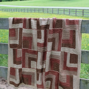 May include: A crocheted blanket with a patchwork design in shades of brown, beige, and red. The blanket is hanging on a wooden fence against a backdrop of green trees and a grassy field.