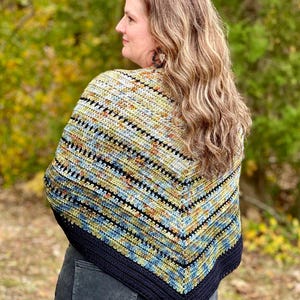 May include: A handmade crochet shawl with a striped pattern in blue, yellow, and black. The shawl is worn over the shoulders and has a dark blue border. The woman is wearing the shawl outdoors. The shawl is a cozy accessory.