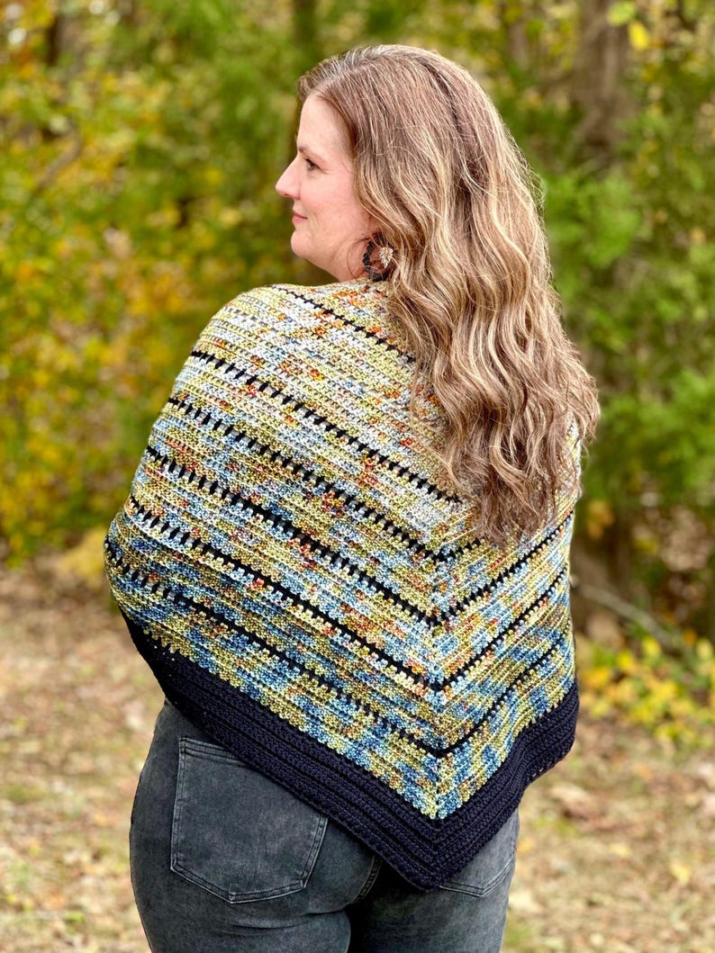 May include: A handmade crochet shawl with a striped pattern in blue, yellow, and black. The shawl is worn over the shoulders and has a dark blue border. The woman is wearing the shawl outdoors. The shawl is a cozy accessory.