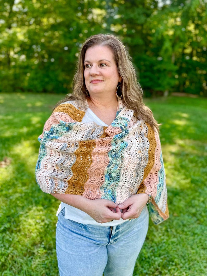 May include: A crochet shawl featuring a wavy, openwork stitch pattern in a variegated colour palette of mustard yellow, soft peach, and speckled blue-green. The rectangular wrap is draped over the shoulders, showcasing a textured yarn construction.