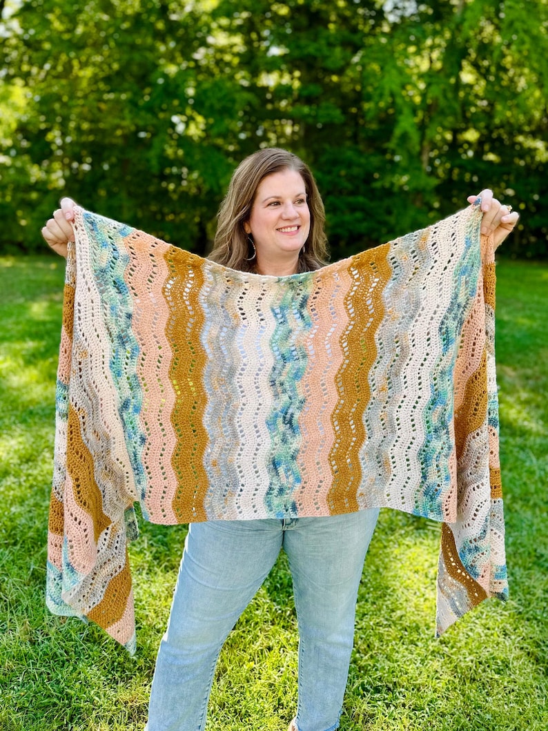 May include: A rectangular crochet shawl featuring a repeating vertical wavy pattern in shades of mustard yellow, soft peach, cream, and variegated teal blue. The textile has an openwork stitch design with visible gaps between the colourful yarn stripes.