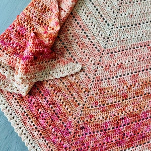 May include: A crocheted blanket with a geometric pattern in shades of pink, orange, and beige. The blanket is folded in half and has a scalloped edge.
