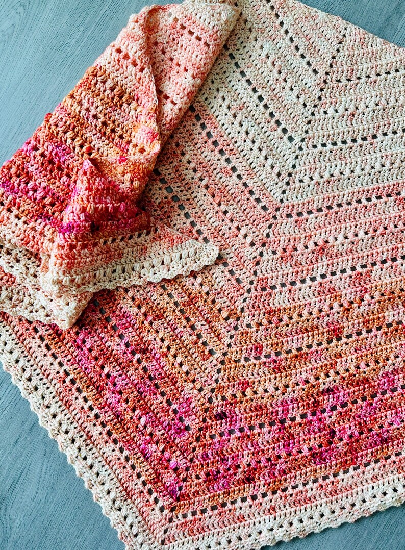 May include: A crocheted blanket with a geometric pattern in shades of pink, orange, and beige. The blanket is folded in half and has a scalloped edge.