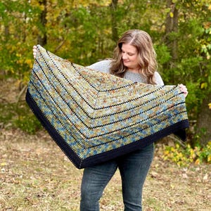 May include: A handmade, colourful crochet shawl with a dark navy border. The shawl features a geometric pattern with shades of blue, green, and tan. The shawl is held up in front of a natural outdoor background.