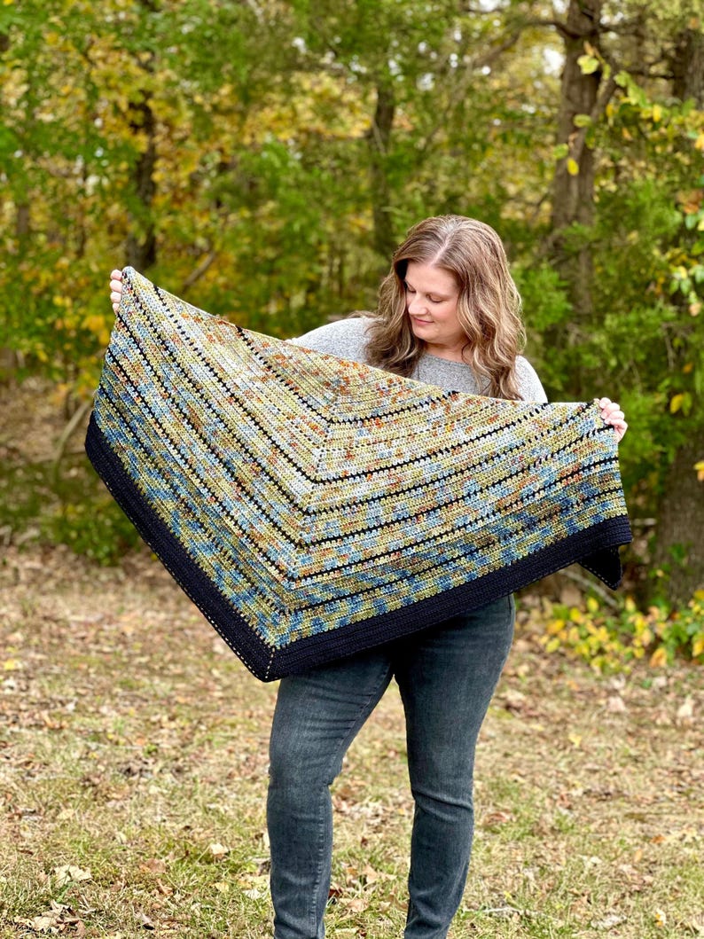May include: A handmade, colourful crochet shawl with a dark navy border. The shawl features a geometric pattern with shades of blue, green, and tan. The shawl is held up in front of a natural outdoor background.