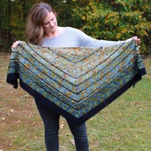 May include: A colourful, triangular shawl with a black border. The shawl features a pattern with shades of blue, green, and orange. The shawl is held up, showcasing its size and design. The background is a natural outdoor setting with trees and grass.