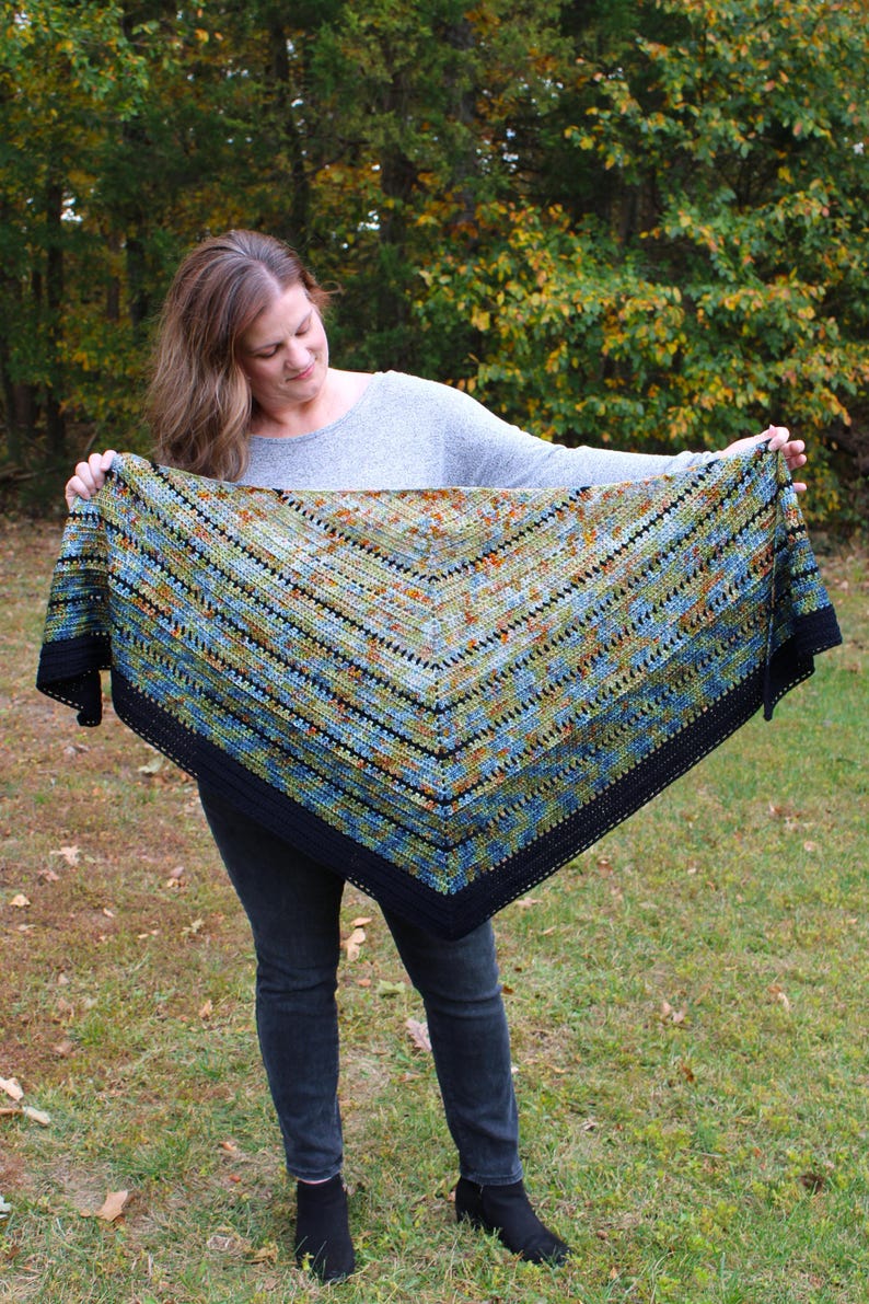 May include: A colourful, triangular shawl with a black border. The shawl features a pattern with shades of blue, green, and orange. The shawl is held up, showcasing its size and design. The background is a natural outdoor setting with trees and grass.