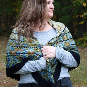 May include: A handmade crochet shawl with a black border, featuring a mix of blue, green, and orange colours. The person is wearing a grey long-sleeved top and black jeans. The background is a blurred view of trees.