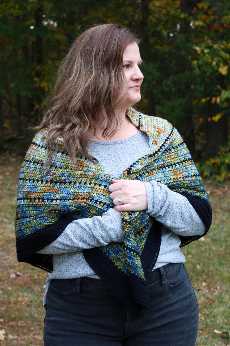 May include: A handmade crochet shawl with a black border, featuring a mix of blue, green, and orange colours. The person is wearing a grey long-sleeved top and black jeans. The background is a blurred view of trees.
