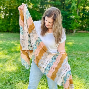 May include: A rectangular crochet shawl featuring a wavy, chevron-inspired pattern. The textile displays a multi-coloured palette of mustard yellow, soft pink, light blue, and cream, with an openwork stitch design throughout the fabric.