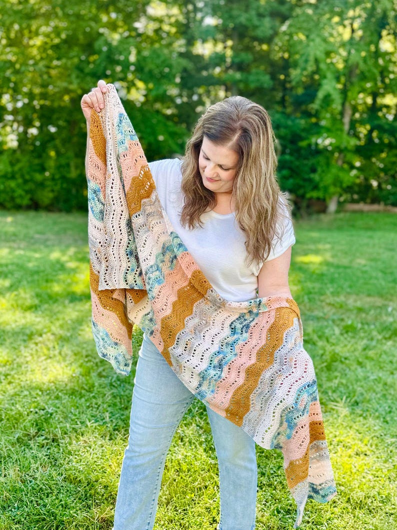 May include: A rectangular crochet shawl featuring a wavy, chevron-inspired pattern. The textile displays a multi-coloured palette of mustard yellow, soft pink, light blue, and cream, with an openwork stitch design throughout the fabric.