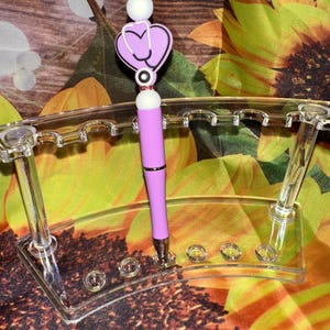 May include: A lavender pen with a stethoscope heart charm and white beads. The pen is displayed in a clear acrylic pen holder. The background features a sunflower pattern.