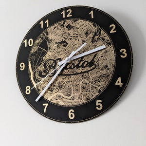 May include: A black and gold clock with a map of Bristol, England as the background. The clock face has gold numbers and white hands.