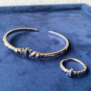 Könnte beinhalten: Ein silberfarbenes Armreif- und Ring-Set. Das Armband ist offen mit einem floralen Design auf der Vorderseite. Der passende Ring hat ein ähnliches florales Design. Beide Teile liegen auf einer blauen Oberfläche.